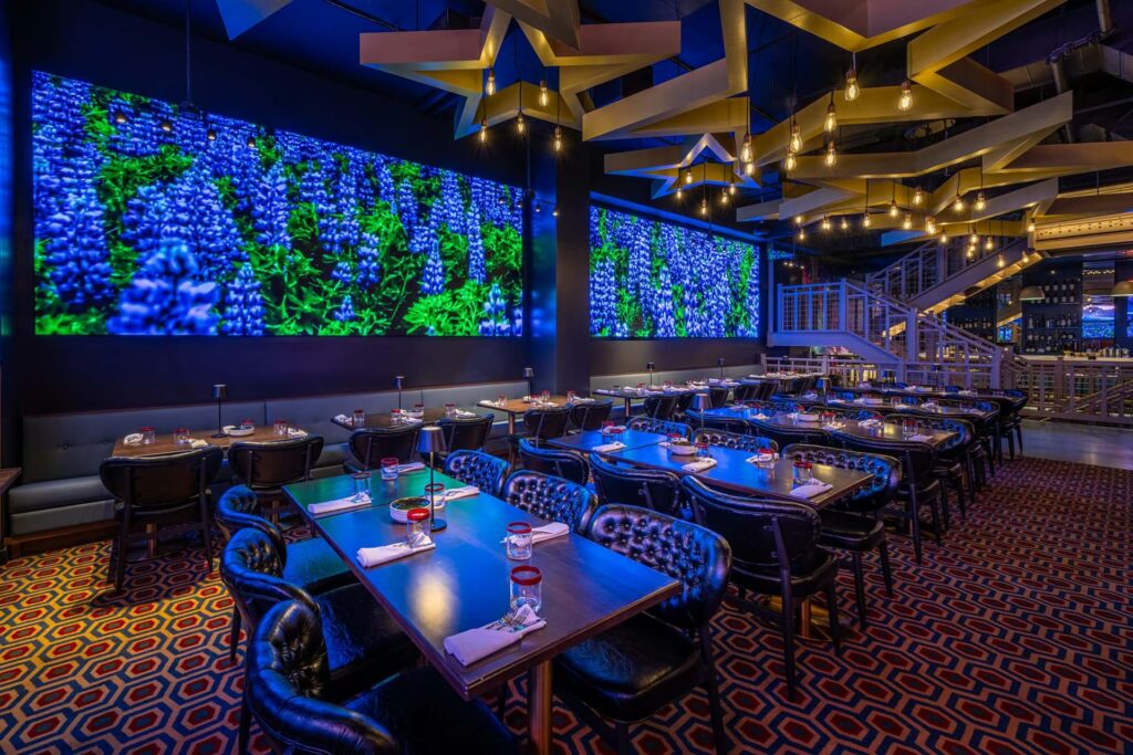 A dramatic dining room in PHNYC with star-shaped light fixtures and large wall screens displaying a vibrant field of blue and purple flowers.