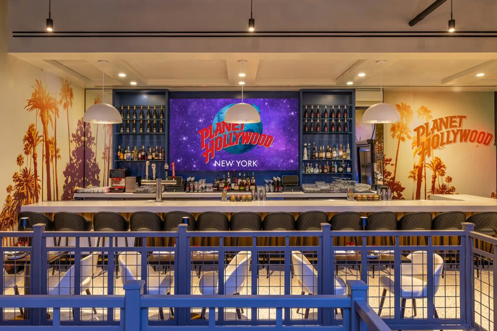 A bright bar in PHNYC featuring a screen with the Planet Hollywood logo against a space backdrop, flanked by shelves and palm tree murals.
