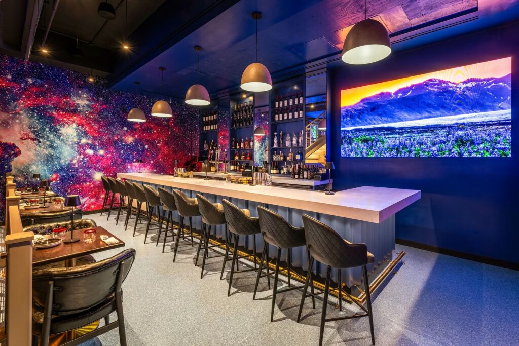 A modern bar area in PHNYC with barstools, a large screen displaying a mountain field, and a dramatic galaxy-print wallpaper on the adjacent wall.