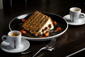 A slice of layered carrot cake, garnished with pecans and carrot curls, is served with two cups of espresso on a dark table at PHNYC.