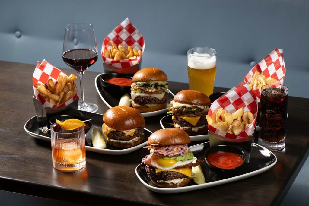 A table setting with four gourmet burgers, two servings of fries, and cocktails, red wine, and beer.