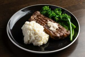 A New York Strip Steak topped with compound butter is served on a black plate with a large scoop of mashed potatoes and steamed broccolini.