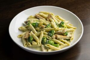 A white bowl filled with penne pasta, sliced chicken, and broccoli florets tossed in a light, creamy sauce.