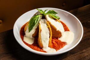 A white bowl holds two breaded and fried lasagna slices covered in white sauce and fresh basil leaves, resting on a bed of red tomato sauce.