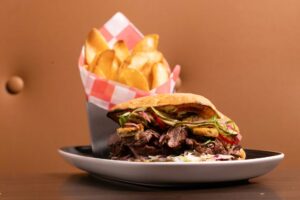 A plate holds a hearty steak sandwich topped with toppings and a side container of potato wedge fries.