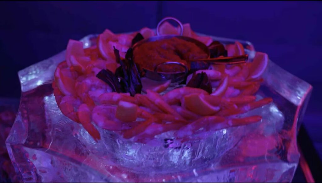 A large shrimp cocktail with lemons and a dipping sauce bowl is served on an ice bowl, illuminated by blue and pink event lighting at PHNYC.