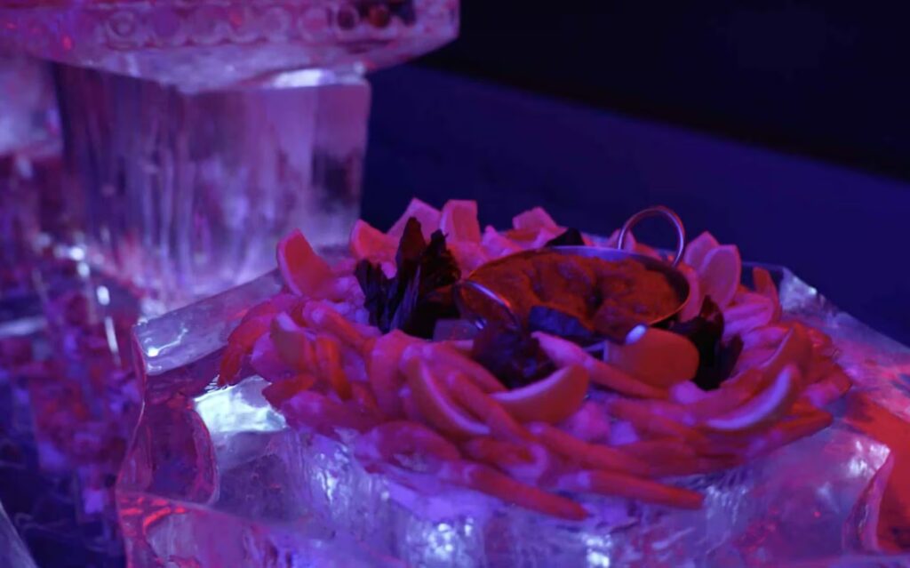 A close-up of a shrimp cocktail and lemon slices resting on a carved ice platter under red and blue event lighting at PHNYC.