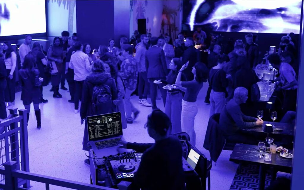 A DJ plays music in the foreground, overlooking a crowded Planet Hollywood NYC event space bathed in purple light, with large video screens on the walls.