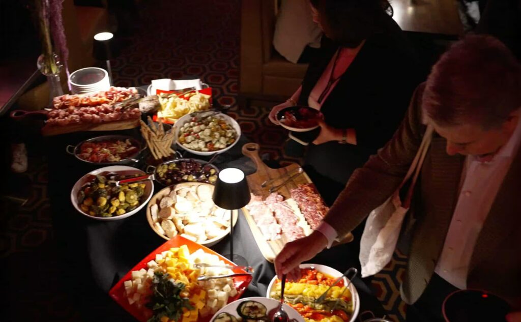 A close-up view of a dimly lit VIP event buffet table loaded with charcuterie, antipasto, cheeses, and salads, with two guests reaching to serve themselves.