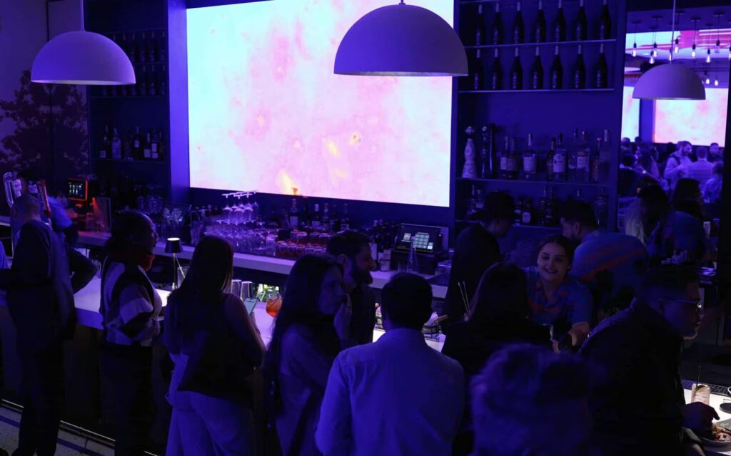 Guests gather at a brightly lit bar in Planet Hollywood NYC, featuring a large screen displaying pink and white abstract art and shelves stocked with bottles.