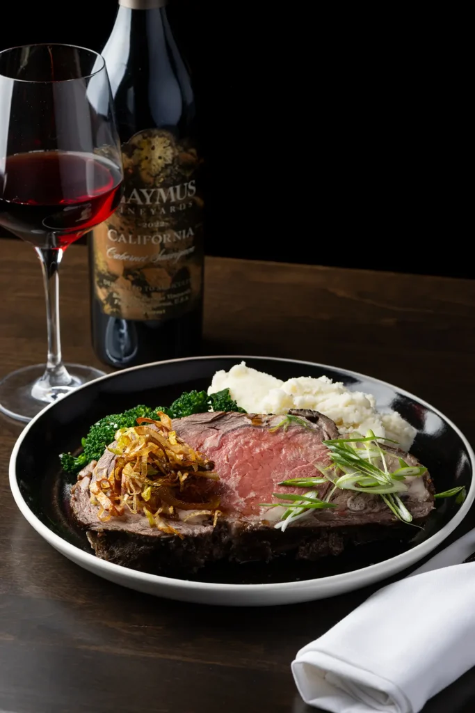 Prime rib dinner with mashed potatoes, broccoli, and a glass of red wine on a table.