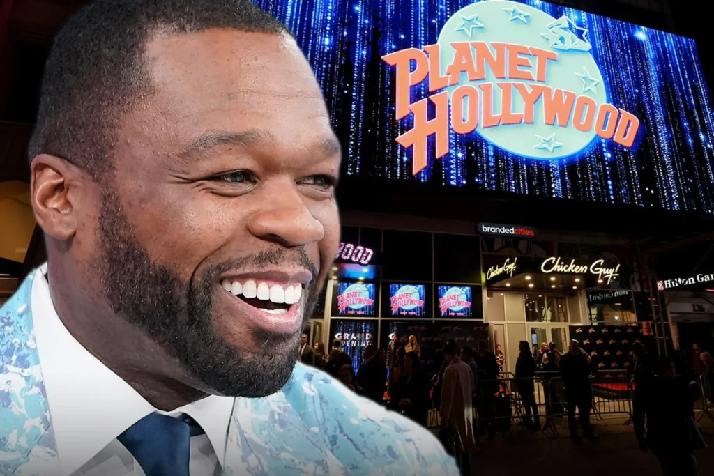 Close-up of 50 Cent smiling in a light blue patterned suit and tie, standing in front of the brightly lit Planet Hollywood sign and entrance in New York City, with a crowd gathered below.
